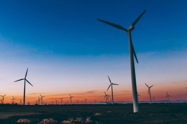 Mavi günbatımı gökyüzüne karşı kırsal tarım ortamında Eko kaynaklardan alternatif temiz yeşil güç üreten pervaneler ile yel değirmenleri İstasyonu silueti. Elektrik üreten türbinlerin çiftliği