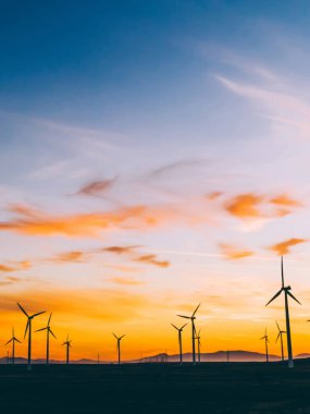 Mavi günbatımı gökyüzüne karşı kırsal tarım ortamında Eko kaynaklardan alternatif temiz yeşil güç üreten pervaneler ile yel değirmenleri İstasyonu silueti. Elektrik üreten türbinlerin çiftliği