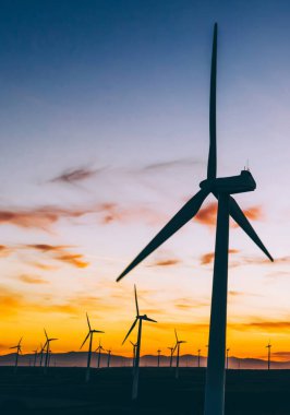 Mavi günbatımı gökyüzüne karşı kırsal tarım ortamında Eko kaynaklardan alternatif temiz yeşil güç üreten pervaneler ile yel değirmenleri İstasyonu silueti. Elektrik üreten türbinlerin çiftliği