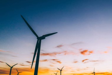 Mavi günbatımı gökyüzüne karşı kırsal tarım ortamında Eko kaynaklardan alternatif temiz yeşil güç üreten pervaneler ile yel değirmenleri İstasyonu silueti. Elektrik üreten türbinlerin çiftliği