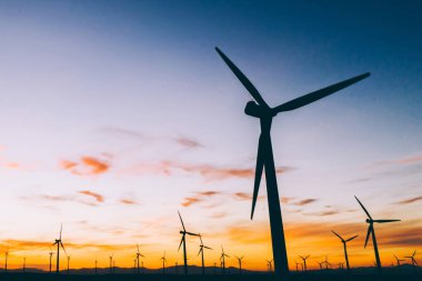 Mavi günbatımı gökyüzüne karşı kırsal tarım ortamında Eko kaynaklardan alternatif temiz yeşil güç üreten pervaneler ile yel değirmenleri İstasyonu silueti. Elektrik üreten türbinlerin çiftliği