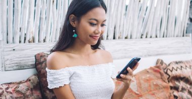 Genç, güzel Asyalı bir bayan, Bali 'de kafenin terasında gülümserken ve akıllı telefon kullanırken, masada cam bir içecekle oturuyor.