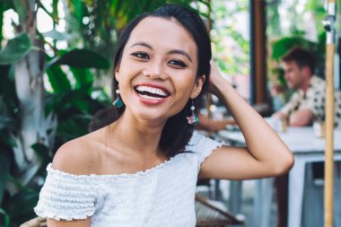 Heyecanlı gülümseyen genç Asyalı kadın, kafeterya terasında bir fincan kahveyle masada otururken, omuzları açık bir şekilde kitap tutuyor.