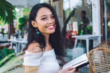 Beyaz omuzlu bluz giyen, açık terasta bir fincan kahveyle oturmuş kitap okuyan neşeli, güzel Asyalı kadının yan görüntüsü.