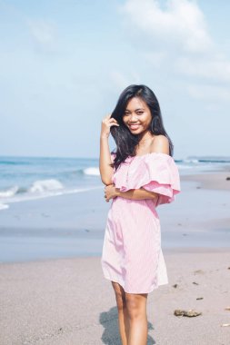 Pembe elbiseli, kumlu sahil şeridinde güneş ışığı altında gülümseyen ve saçlarına dokunan romantik etnik kadın Bali.