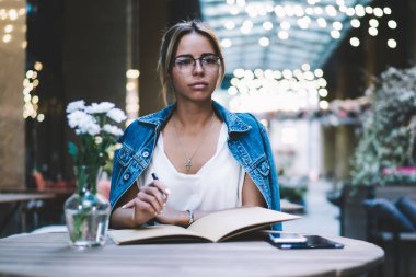 Optik gözlükler içinde düşünceli hipster kız durulıyor ve kafeteryada otodidakt hakkında düşünme iken, eğitim için ders kitabı ile masada oturan düşünce genç kadın yazar