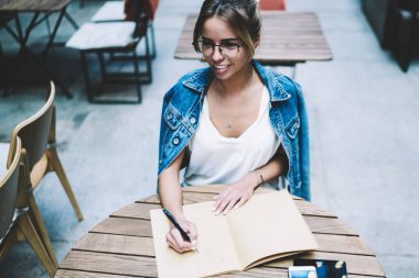 Keyifli kadın yazar, kafeterya masasında eğitim için Notebook ile oturan heyecanlı bir fikir ile kompozisyon yaratma, göz giyim çekici gülümseyen hipster kız otodidakt kapalı ücretsiz zaman zevk