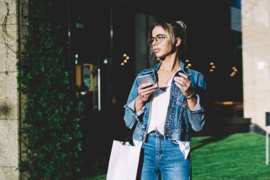 Şaşkın kadın elinde alışveriş çantası ve akıllı telefon tutarak ve güneşli bahar gününde uzağa bakarak, reklam alanında ayakta iken Denim kıyafeti giymiş çekici hipster kız arama bekliyor