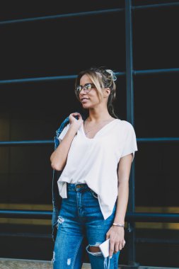 Elle akıllı telefon gadget ile kentsel ayarı arkadaş beklerken uzak görünümlü Casual Denim kıyafeti giyinmiş genç pozitif hipster kız, açık havada trendy optik gözlükler mutlu gülümseyen kadın