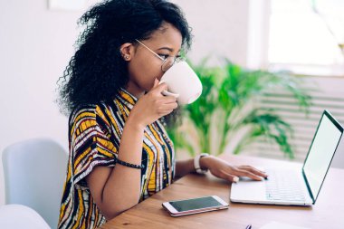 Yetişkin, modern, gündelik kıyafetli, gözlüklü, dizüstü bilgisayarlı, evde çay içerken boş ekranlı bir masada oturan bir kadının yan görüntüsü.