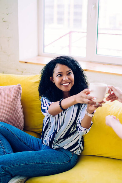 From above African American woman with smile glad to get white cut from hand of young colleague during work with laptop sitting on yellow sofa