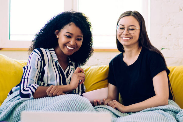 Smiling African American woman with diverse female friend sitting on yellow sofa under blue cozy plaid and watching comedy movie on laptop