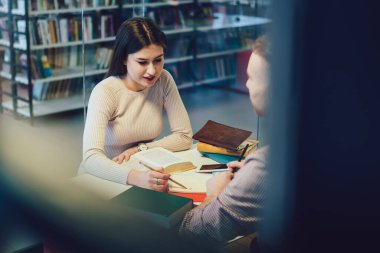 Modern kütüphanede cam odada otururken erkek sınıf arkadaşına ödevlerini açıklayan genç bir kadın.