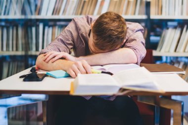 Yorgun erkek girişimci resmi takım elbise giyip kütüphanede kitapların yanında defter ve kitaplarla yüz üstü yatıyor.