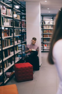 Odaklanmış ve meşgul bir erkek öğrenci kütüphanede kitaplıkların arasında otururken kadınlar sayfalarca kitap okuyup ilginç hikayeler okuyorlar.