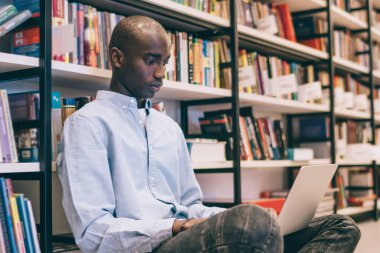 Günlük giysiler içinde kel bir adam kütüphanede yerde oturuyor ve taşınabilir dizüstü bilgisayarla tek başına kişisel proje yapıyor.