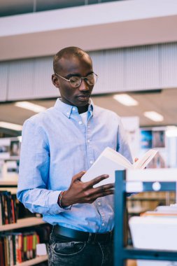 Modern kütüphanede kitap standının yanında duran, gömlek ve gözlüklü, çalışkan Afrikalı Amerikalı genç adam.