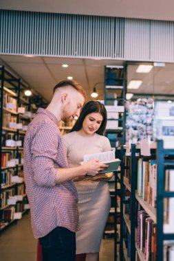 Modern kütüphanede kitapçıların yanında dururken gündelik kıyafetlerle kitap çeviren çok kültürlü gençlerin yan görünüşü
