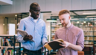 Gömlekli ve siyah kotlu çok ırklı erkekler kitaplıkların yanında durmuş edebiyat hakkında konuşuyor ve üniversite kütüphanesine bakıyor.