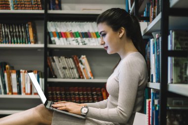 Kitap raflarına yaslanıp kütüphanede yerde otururken dizüstü bilgisayar kullanan düşünceli, konsantre bir kadın.