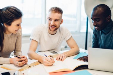 Modern kütüphanede kitaplarla ve dizüstü bilgisayarlarla masada otururken, günlük giysiler içinde neşeli çoklu etnik gençler deftere yazı yazıyorlar.