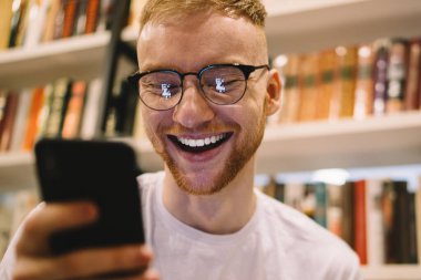 Beyaz tişörtlü ve gözlüklü neşeli yetişkin bir öğrenciden gülümseyen ve ekrana odaklanan modern kütüphanede kitaplığın yanında akıllı telefon kullanan.