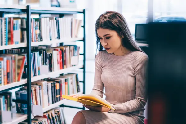Moda elbiseli genç bayan kitaplığın yanında oturuyor ve üniversitenin hafif kütüphanesinde ödevlerini yaparken ilginç kitaplar okuyor.