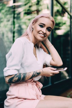 20 yaşındaki mutlu bir kadının portresi modern cep telefonuyla eğleniyor dışarıda kendini iyi hissediyor, dövmeli hippi kız elinde akıllı telefon cihazıyla elinde 4 g kablosuz ve gülümsüyor.