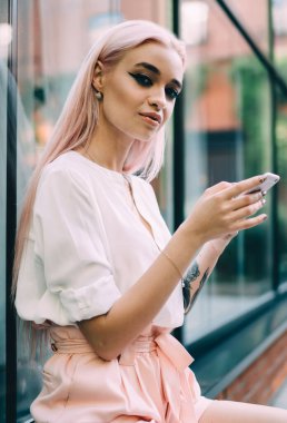 Elinde dijital teknolojisi olan çekici bir hipster kızın portresi açık havada oturuyor ve poz veriyor, güzel beyaz kadın blogcu pembe saçlı ve cep telefonu cihazıyla kameraya bakıyor.