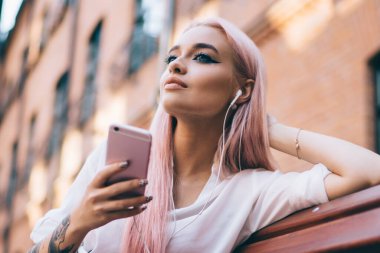 Kulaklık takan güzel genç bir kadın şehir bankında otururken podcast 'ten keyif alıyor. Pembe saçlı, cep telefonundan gelen ses kitabına odaklanmış beyaz bir meloman.