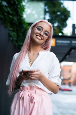 Moda kıyafet kavramı ve elinde cep telefonu cihazıyla şehir atetinde poz veren şık milenyum insanı. Optik gözlüklü heyecanlı hippi kızın yarım boy portresi. Keyfini çıkarıyor.