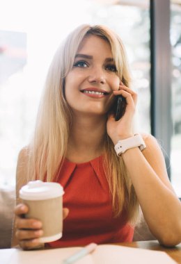 İyimser genç bir kadın elinde sıcak bir bardak içecekle rahat bir restoranda dinlenirken ve akıllı telefondan konuşurken gülümseyip başka tarafa bakıyor.