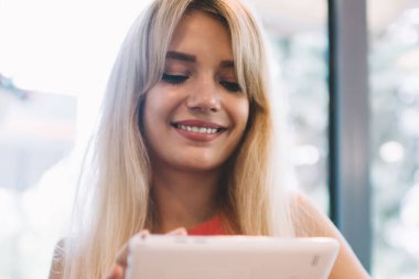 Sevinçli, sarı saçlı, neşeli gülümseyen ve modern tablet kullanan genç bir kadın rahat bir kahvehanenin penceresinin önünde, kanepede oturuyor.