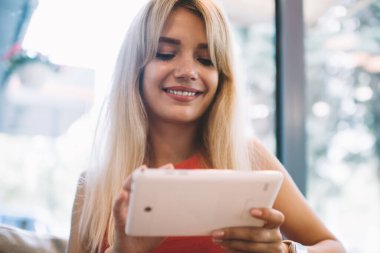 Sevinçli, sarı saçlı, neşeli gülümseyen ve modern tablet kullanan genç bir kadın rahat bir kahvehanenin penceresinin önünde, kanepede oturuyor.