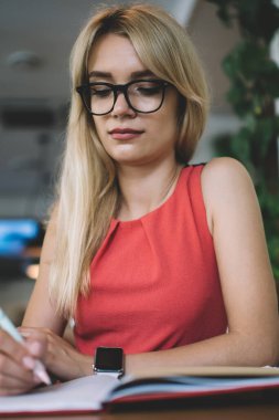 Aşağıdan, gözlüklü, genç, çekici, sarışın bir kadın çalışanın samimi, modern bir kafede otururken notlar ve fikirler alması.