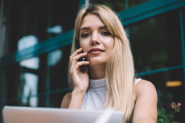 Sarışın, gülümseyen, kolsuz tişörtlü, cep telefonuyla konuşan, şehirdeki bir kafede dizüstü bilgisayarla oturan akıllı kadın.