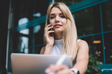 Sarışın, gülümseyen, kolsuz tişörtlü, cep telefonuyla konuşan, şehirdeki bir kafede dizüstü bilgisayarla oturan akıllı kadın.