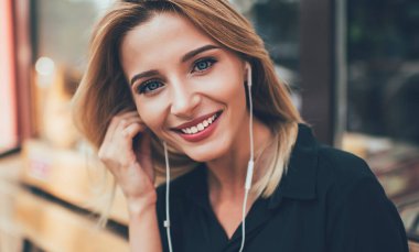 Müzik hobisi için zaman harcarken sevimli gülümseyen ve beyaz dişleriyle kameraya bakan güzel bayan meloman portresini yakından çek. Elektronik kulaklıklı mutlu sarışın kadın dışarıda poz veriyor.