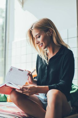 Rahat bir kahve dükkanında oturmuş not defterinde toplantı saatini kontrol eden sıradan giyinmiş, neşeli başarılı bir kadın sınav hazırlıkları için ders kitabından bilgi okuyor.