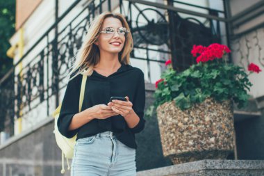 Gündelik giyinmiş neşeli kadın turist ellerinde modern akıllı telefon cihazları ve yaz tatillerinde şehir sokaklarında gezen şık güneş gözlüklü mutlu kadın dışarıda gülümsüyor.