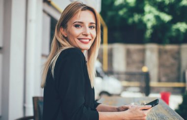 Teknoloji ve nesil kavramı, günlük tişört giymiş başarılı beyaz kadın blog yazarı portresi sokak kafeteryasında elinde dijital aletlerle kameraya gülümsüyor.