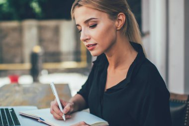 20 yaşındaki zeki beyaz kız öğrencinin öğrenim için dışarıda ders kitabı ve dizüstü bilgisayarla çalışmasının yan görüntüsü. Modern cep telefonu cihazında saati kontrol eden çekici bir kadın.