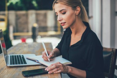 20 yaşındaki zeki beyaz kız öğrencinin öğrenim için dışarıda ders kitabı ve dizüstü bilgisayarla çalışmasının yan görüntüsü. Modern cep telefonu cihazında saati kontrol eden çekici bir kadın.