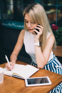 Açık kafede masada otururken ve serbest iş yaparken sarı saçlı cep telefonu konuşurken ve açık dizüstü notları yapma ile çekici kadın