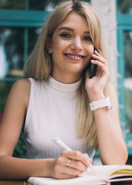 Güzel genç kadın gülümsüyor ve cep telefonu konuşurken kamera bakmak ve açık not defteri içinde masada Not yapma kapalı kafe