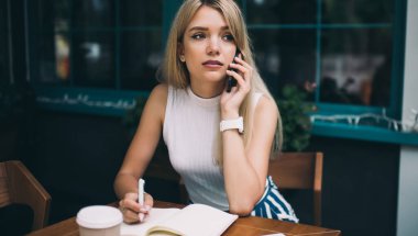 Güzel genç kadın düşünme ve uzak görünümlü cep telefonu ve açık kafede masada not defterinde yazma konuşurken