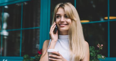 Sıcak içki fincan ile güzel genç kadın gülümseyen ve uzakta açık kafede serbest iş yaparken ve ara sırasında akıllı telefonda konuşan