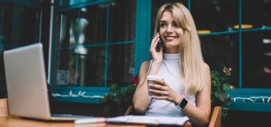 Sıcak içki fincan ile güzel genç kadın gülümseyen ve uzakta açık kafede serbest iş yaparken ve ara sırasında akıllı telefonda konuşan