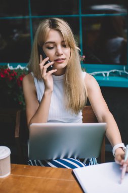 Şehir sokak açık kafede serbest iş yaparken laptop ile akıllı telefon konuşma ve Pano üzerinde notlar yapma sahip güzel genç bayan