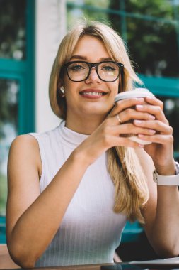 Güzel genç kadın trendy gözlük dostu gülümseyen ve masada otururken ve açık kafede paket sıcak içecek tadını çıkarırken kamera bakıyor